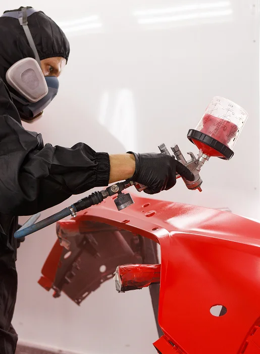 Peintre automobile en tenue de protection appliquant de la peinture rouge sur une pièce de voiture Les Essarts-le-Roi dans les Yvelines 78